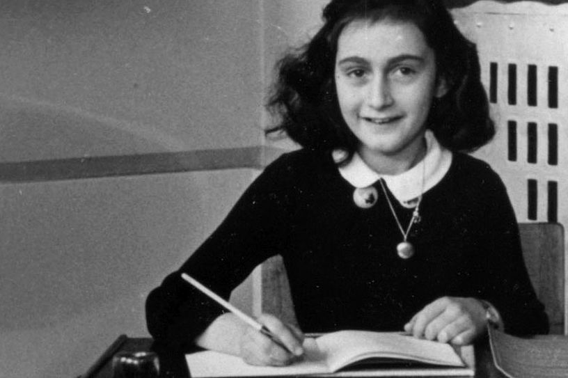 anne frank sitting at a desk and writing in a notebook